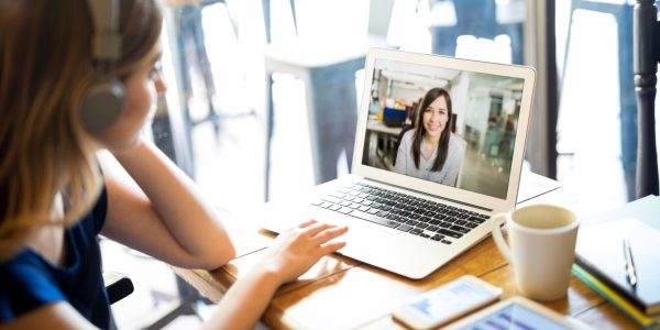 Woman wearing headphones and participating in a video conference call on a laptop while telecommuting from a cafe
